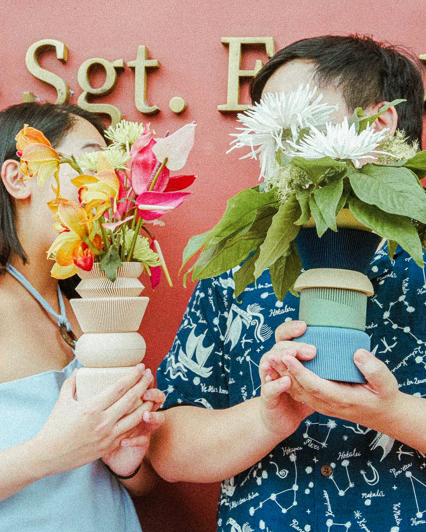 Two people holding flower pots with a pink background